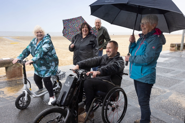 picture for projectStory: A man on a three wheeled wheelchair and a  woman on a seated scooter on a board walk next to the beach smile. Three people  stand behind.