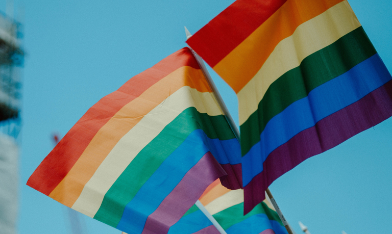 Two pride flags in the wind in front of a clear blue sky.