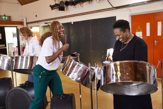 One woman playing steel pans and laughing while another women keeps the beat on a wood block percussion instrument.